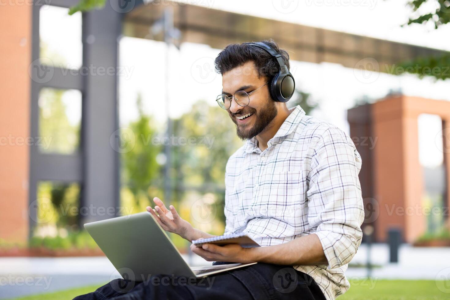 Smiling young Indian man sitting on a bench outside wearing headphones, holding a notebook with a pen and talking on call. photo