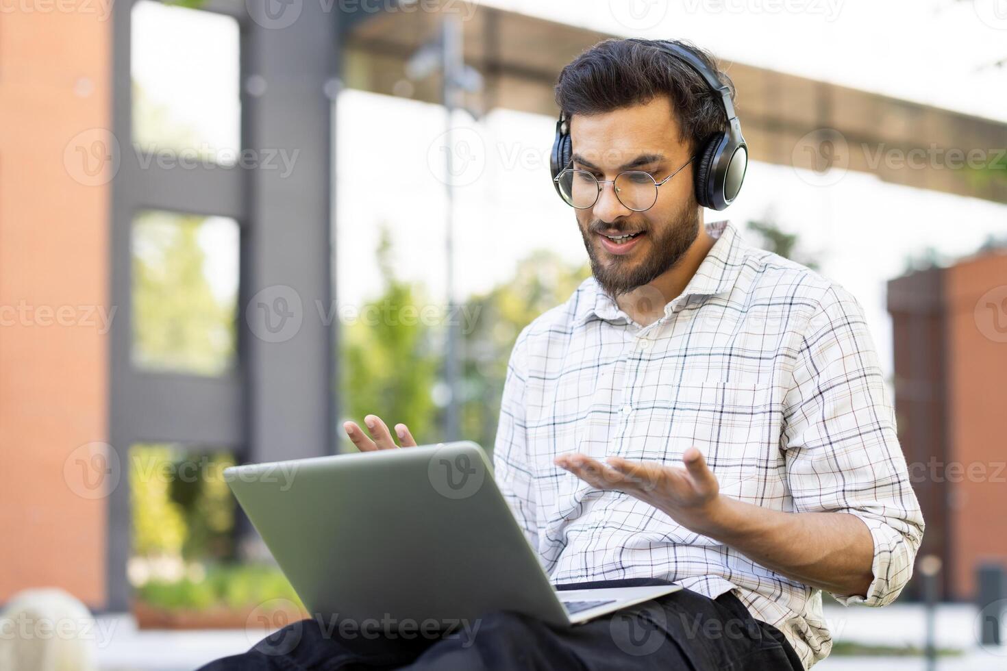 A young student wears headphones and engages in a call outdoors, using his laptop with a friendly expression. photo