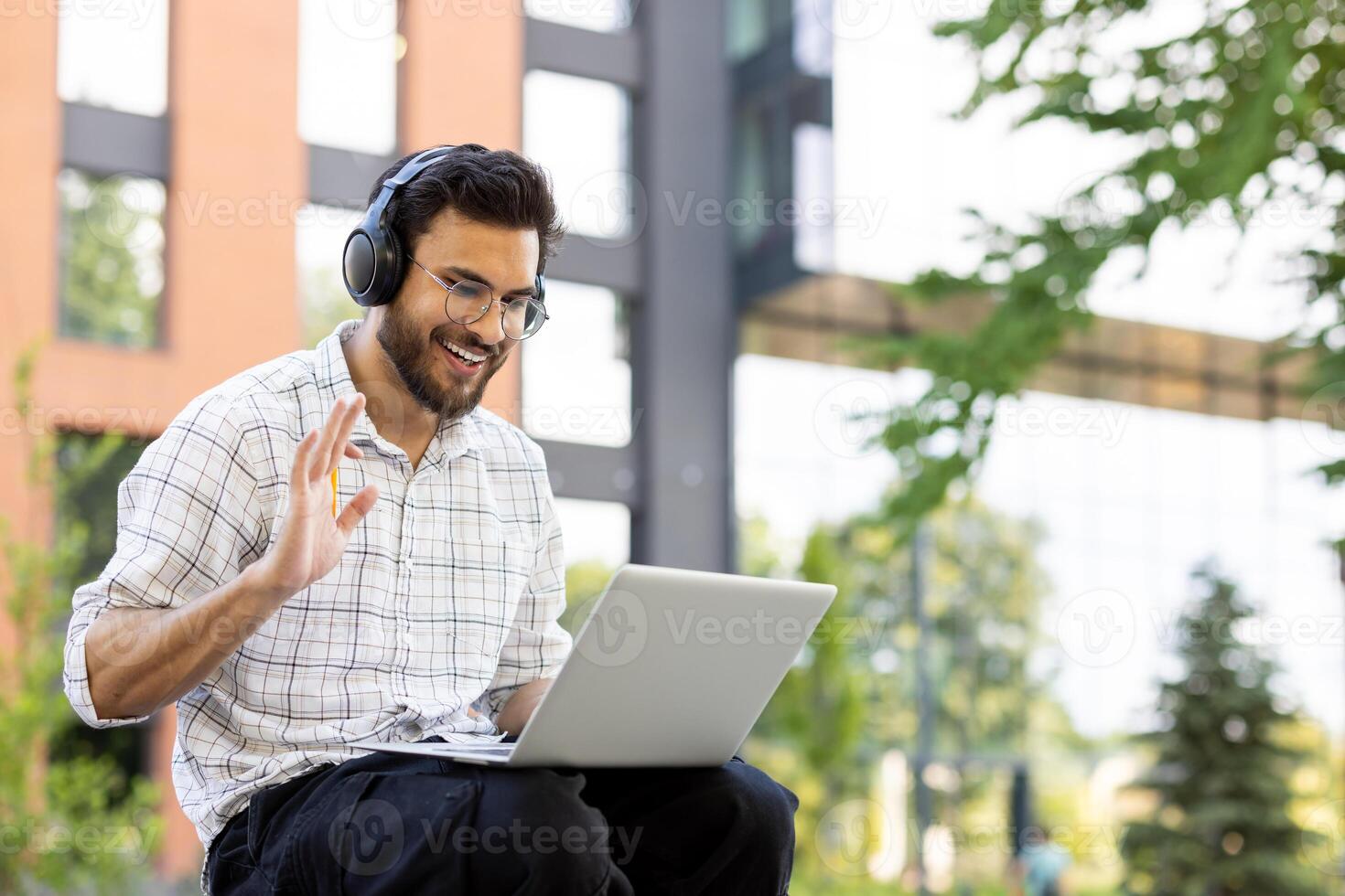 A smiling man with glasses wearing headphones waves while using a laptop outdoors, appearing to be on a call. photo