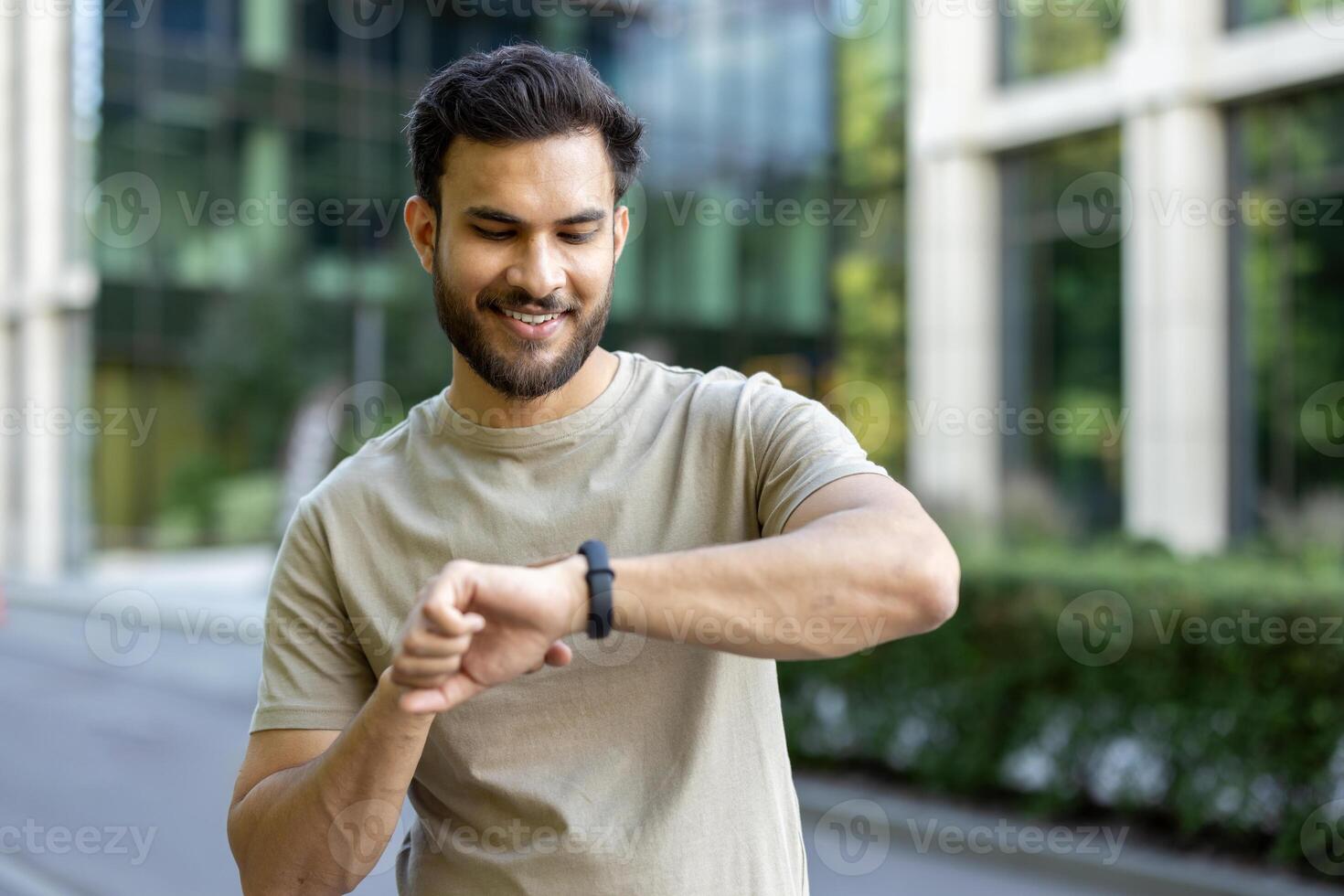 A smiling man outdoors checking his watch, possibly monitoring activity or time. The background shows a modern building and greenery. photo