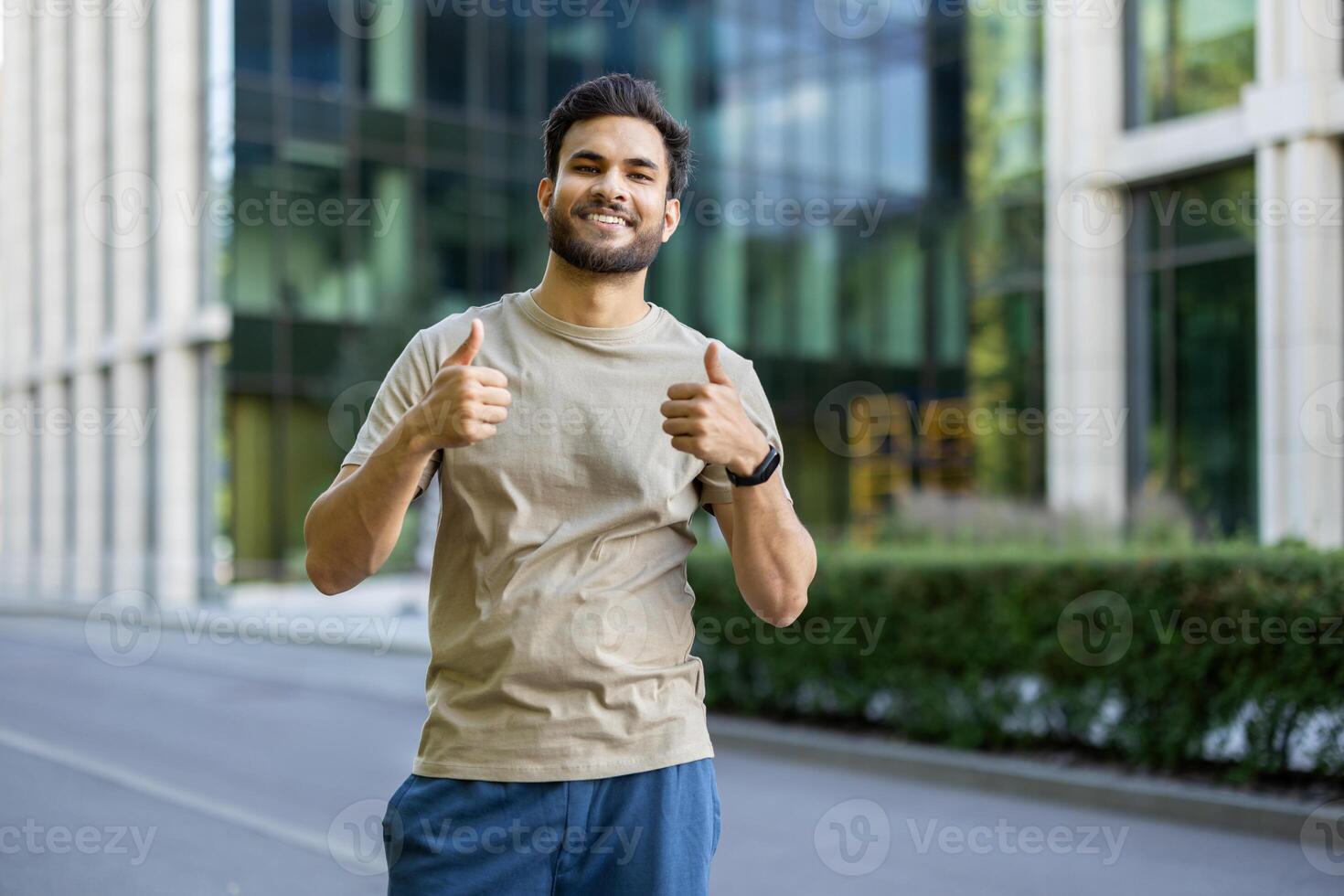 A smiling man gives a thumbs up gesture outdoors. He is likely expressing approval or showing positivity, in a city setting. photo
