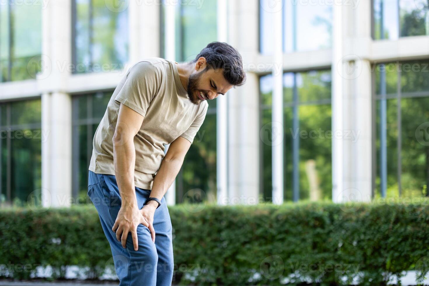 A man, clutching his knee in apparent pain outdoors, seemingly injured during a physical activity. The building is in the background. photo