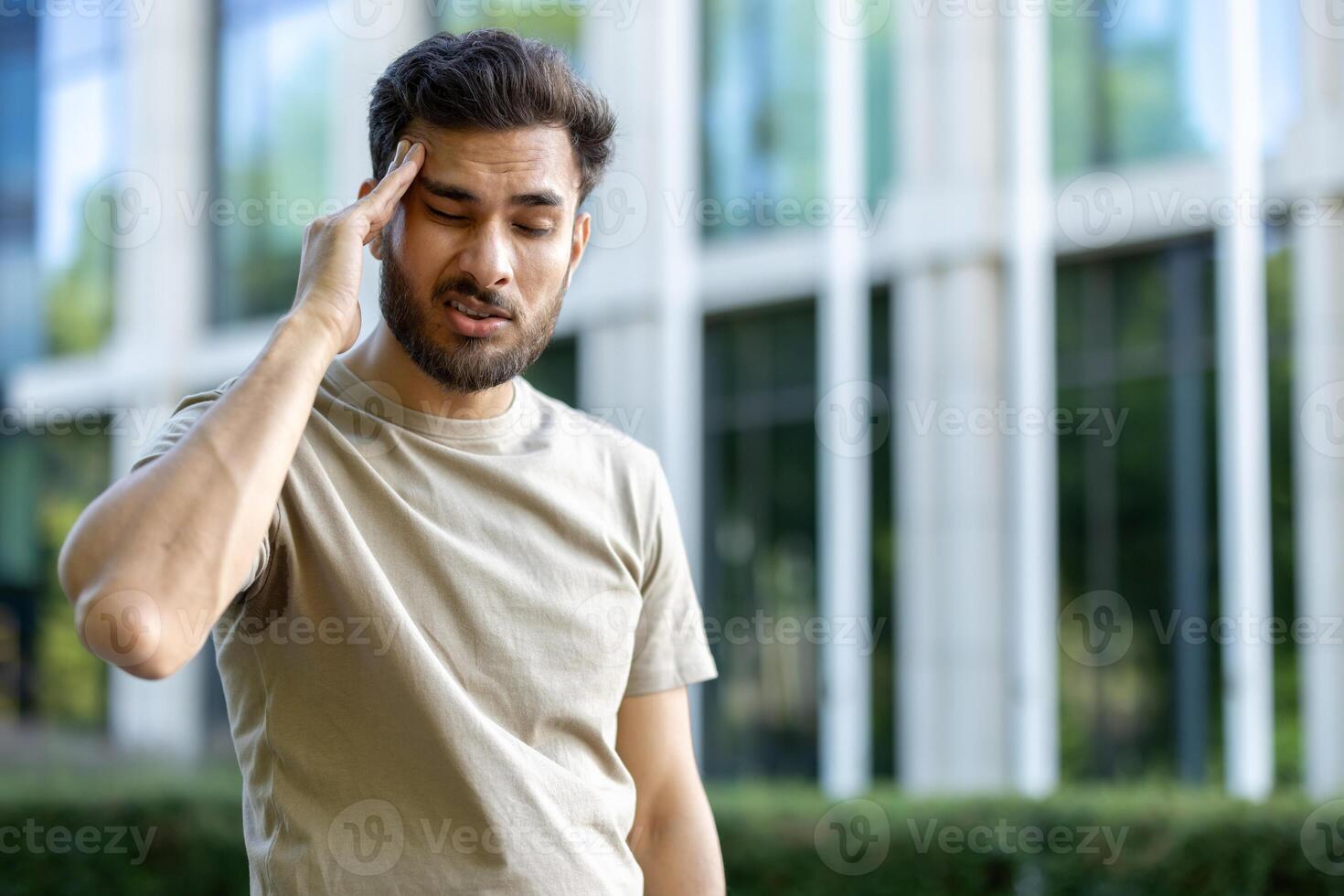 A young man, possibly experiencing headache or discomfort, touches his temple while outside. The background features a blurred modern building. photo