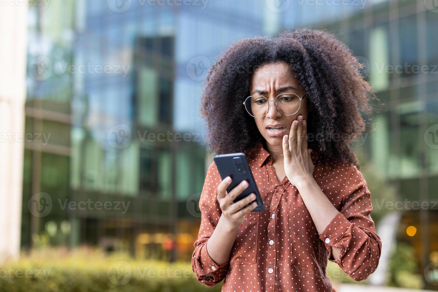 An anxious businesswoman in glasses reacts with concern while looking at her phone in an urban setting. photo