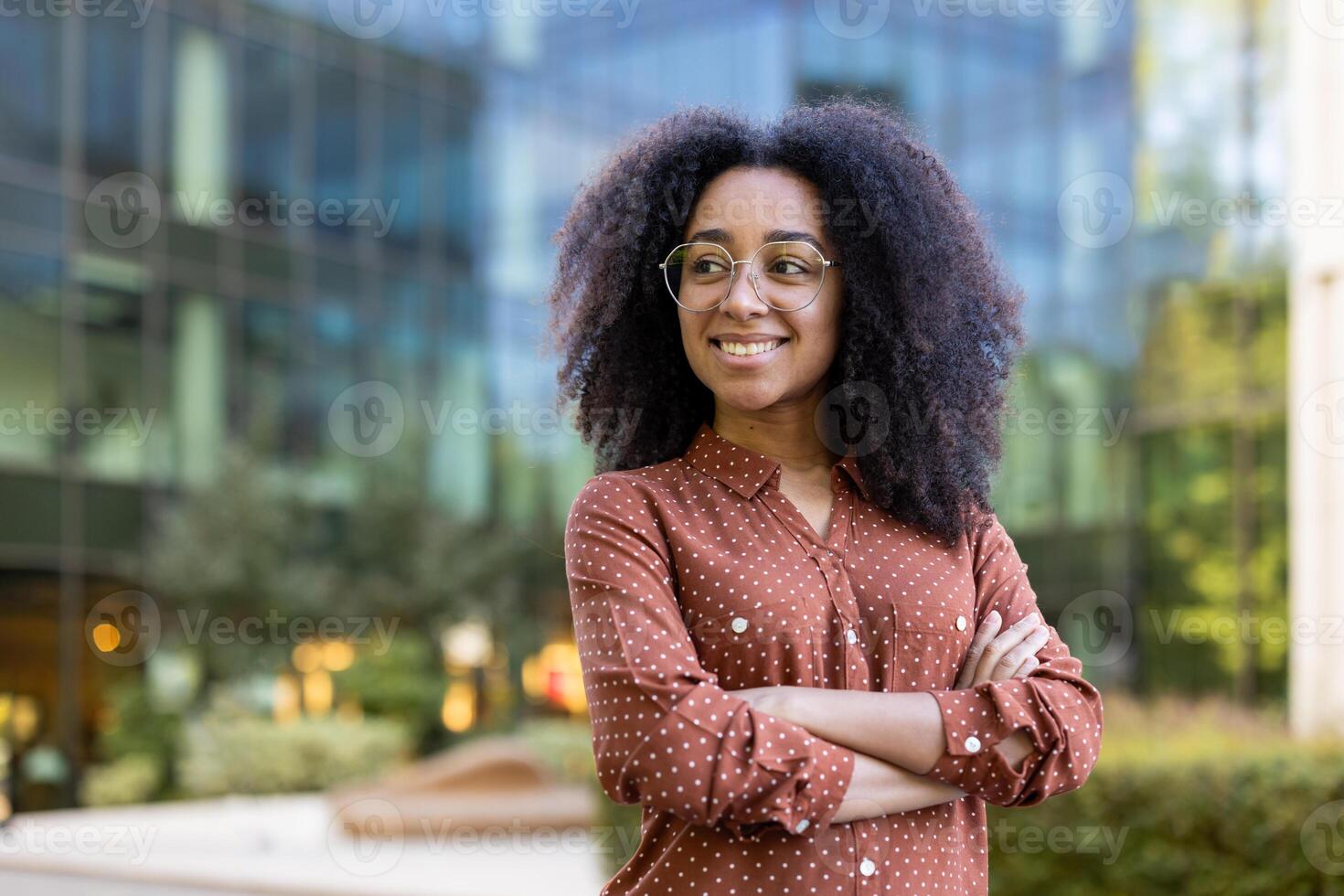 A beautiful, smiling woman with curly hair, wearing glasses and a polka dot shirt, stands with arms crossed in front of a modern building. photo