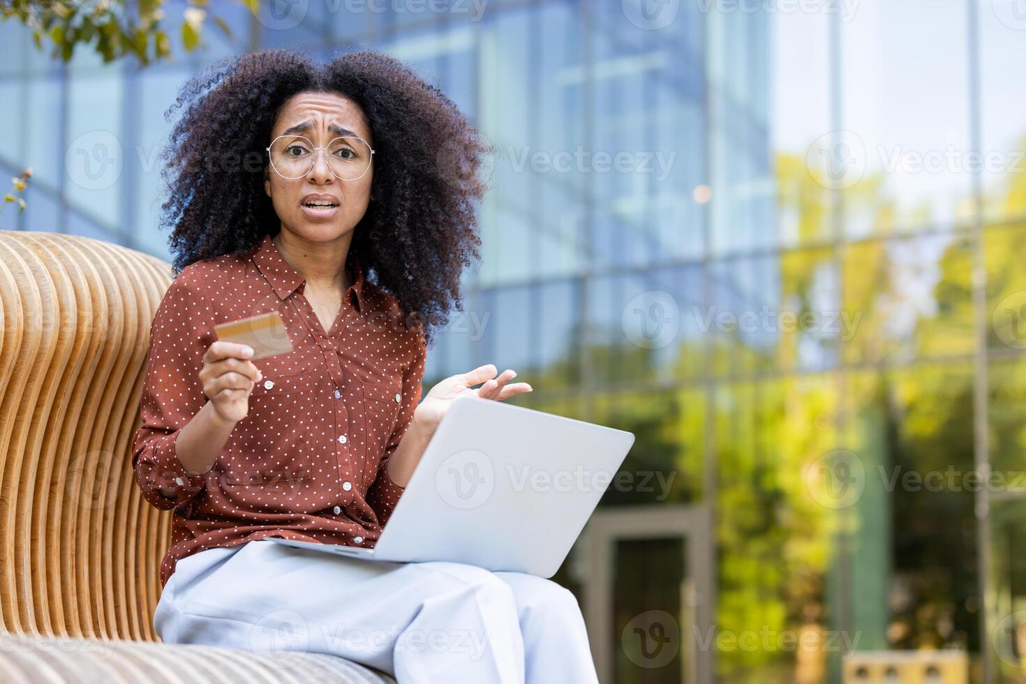 A concerned woman with curly hair holds a credit card and looks at a laptop with a worried expression, possibly experiencing a financial issue. photo