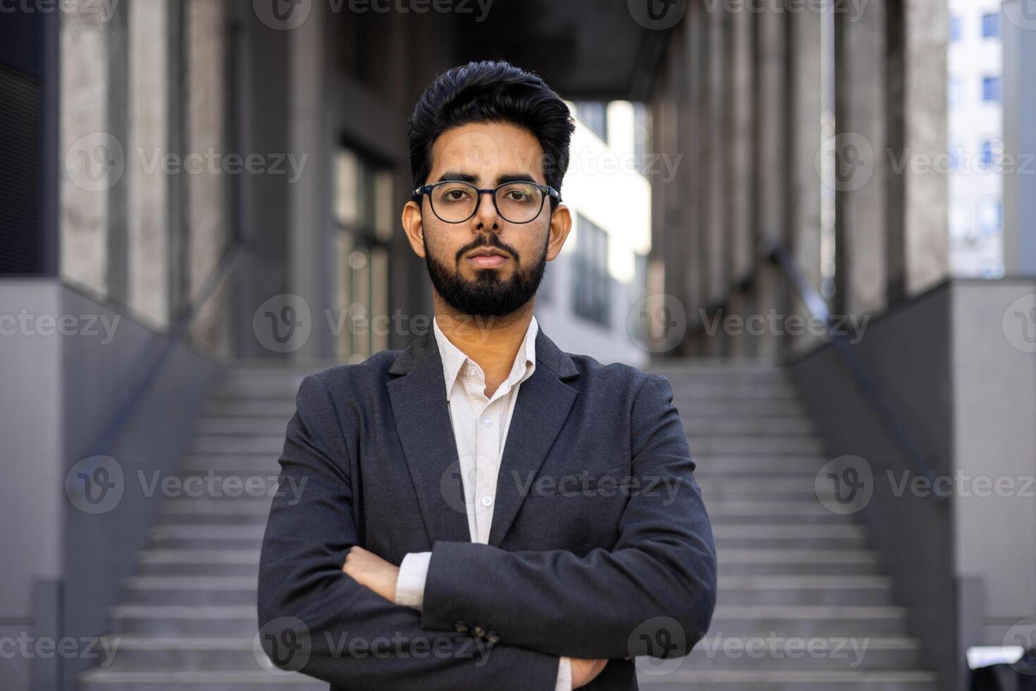 Portrait of serious Hindu businessman standing outside the office with his arms folded on his chest, a strict boss in glasses and a suit looking confidently into the camera. photo