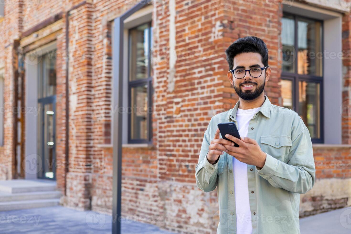 Portrait of young successful student outside university campus, man smiling looking at camera and using app on phone, typing message and browsing social media, Indian man holding phone with backpack. photo