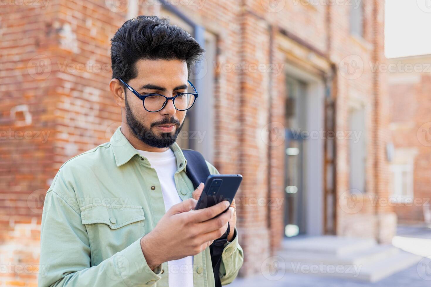 Young indian guy in eyeglasses looking at smartphone screen while standing with backpack outdoors. Focused young man checking online classes schedule for attending courses in university campus. photo