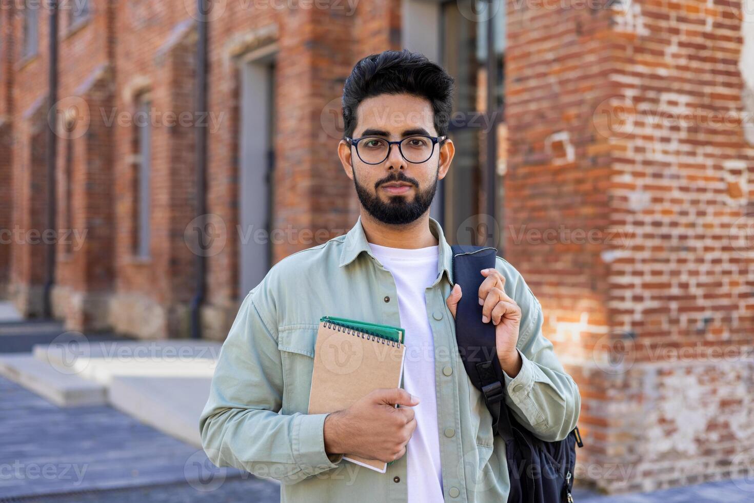 Confident young male Indian student standing outside a university campus with a backpack, holding a notebook and looking seriously at the camera. photo