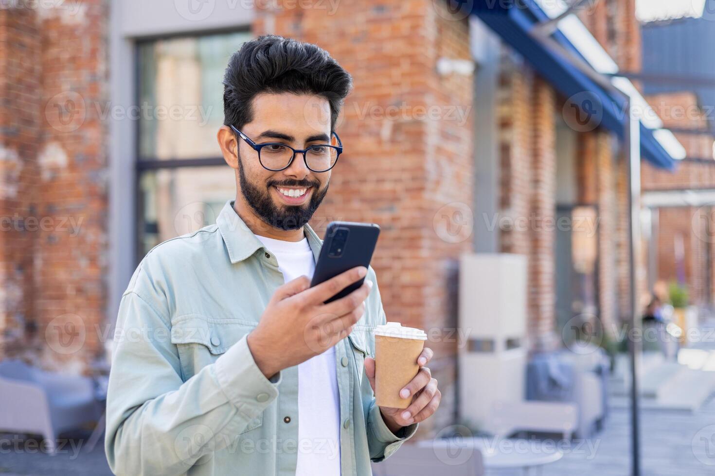 A happy Indian man is standing on the street, holding a cup of coffee, smiling and looking at the smartphone screen. photo