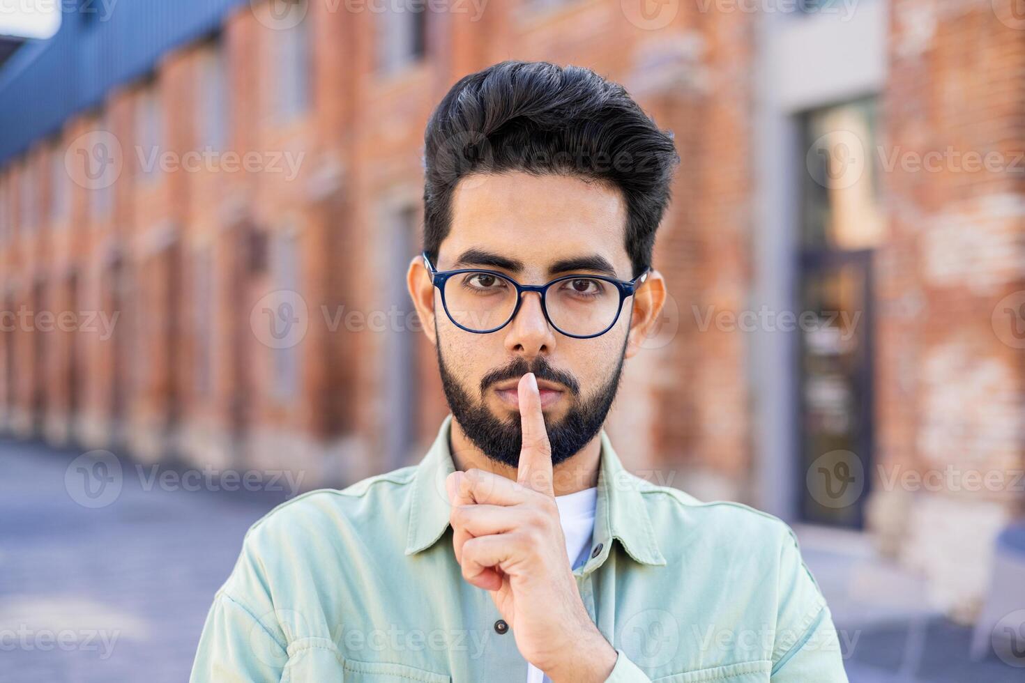A young Indian man stands on the street, puts his index finger to his lips, looks at the camera. photo