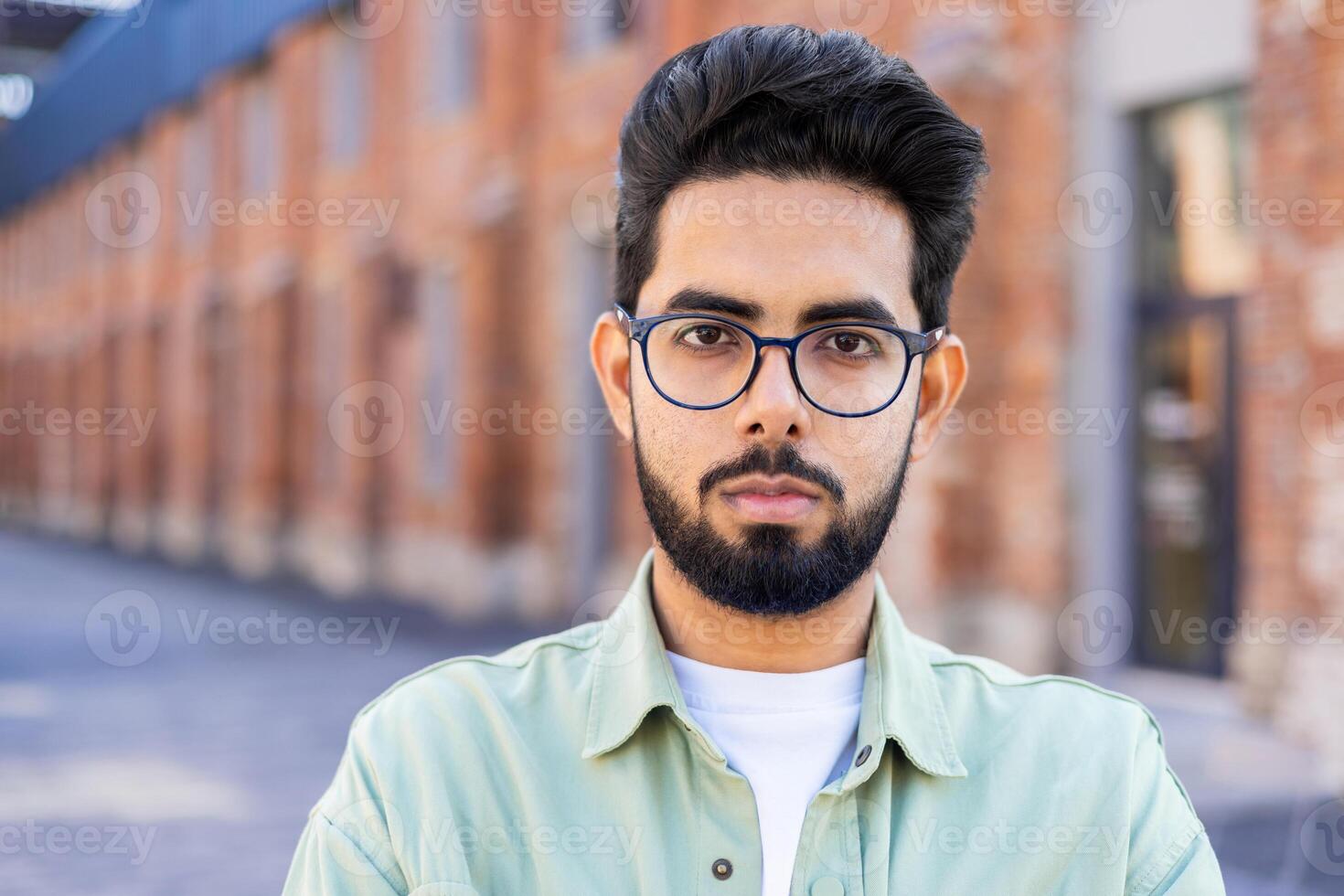 A close-up portrait of a young Indian man standing outside on his forehead and looking seriously into the camera. photo