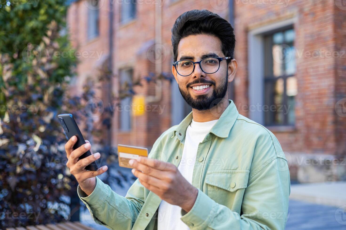 Young arab man holding mobile phone and credit card while looking at camera with wide smile. Cheery bearded guy confirming online cashless payment in banking application during walk outside. photo