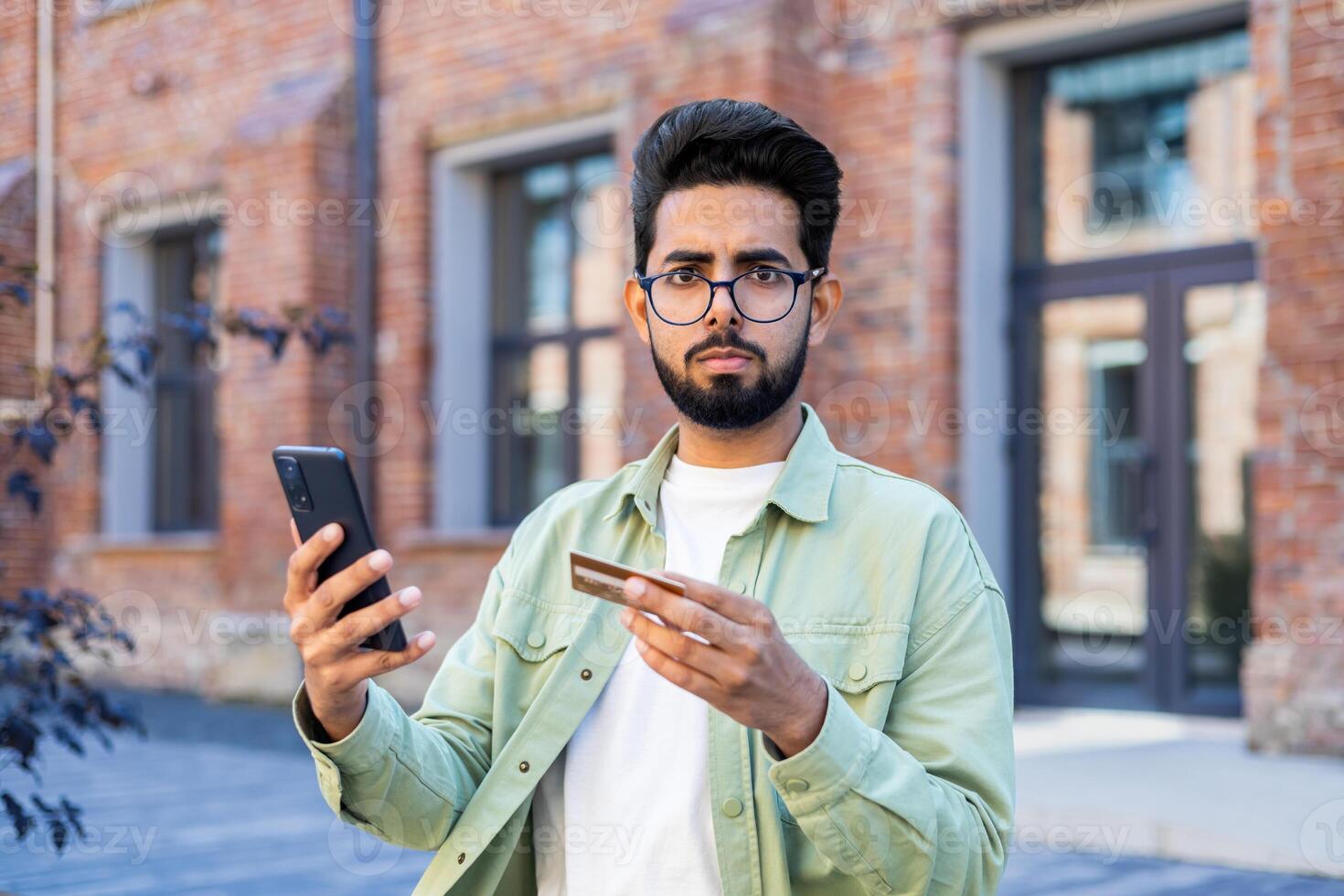 Portrait of a serious young Indian man sitting on a bench outside, holding a mobile phone and a credit card, looking worriedly at the camera. photo