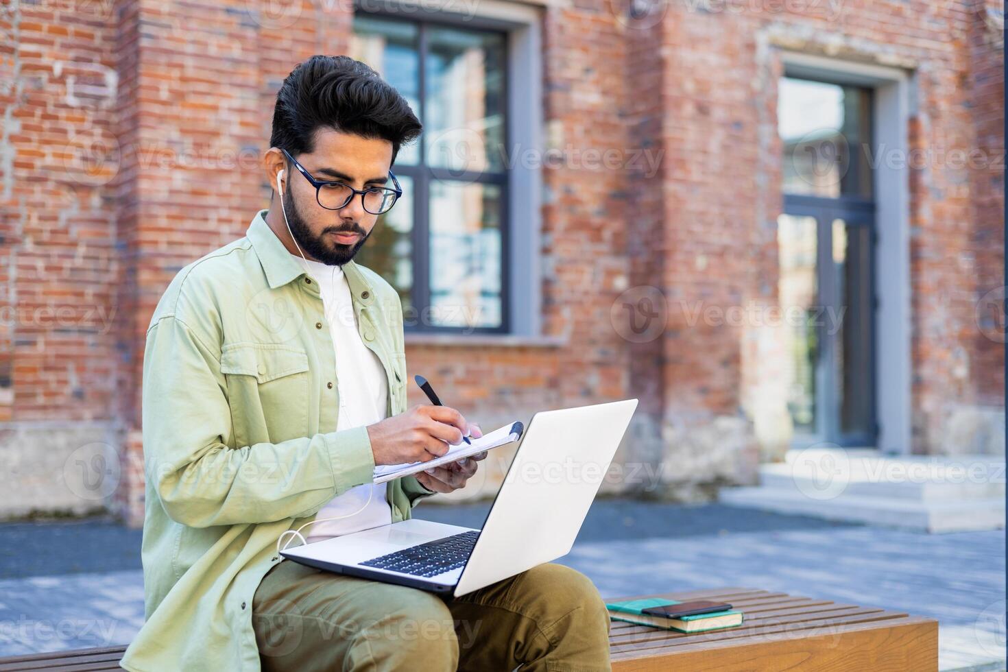 Hindu student in headphones sitting outside on a bench, holding a laptop on his lap, writing in a notebook, studying online. photo