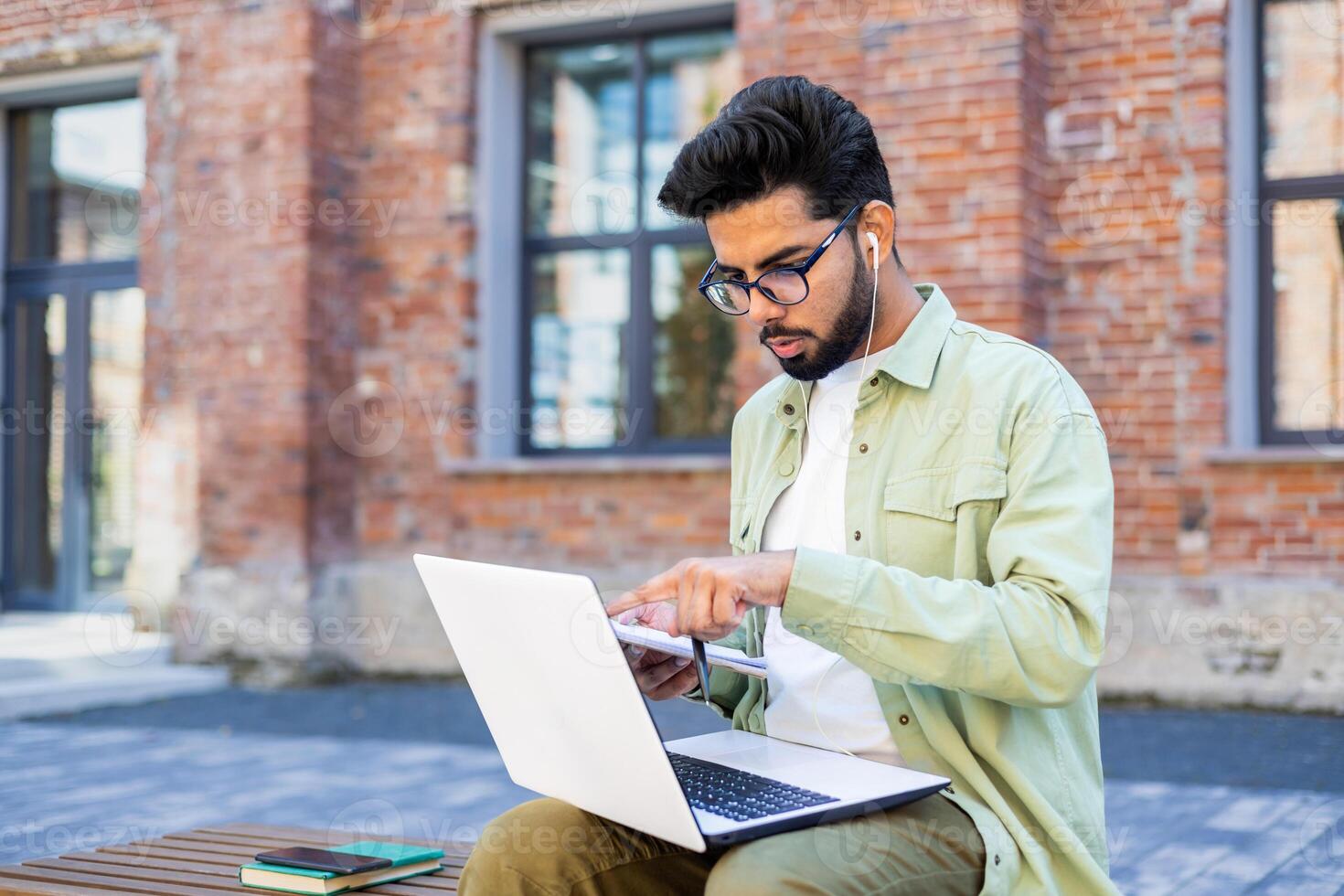 Young indian man in olive shirt sitting on bench at campus with pc on laps and wired earphones. Concentrated student pointing with finger at laptop screen while talking with group mate in online call. photo
