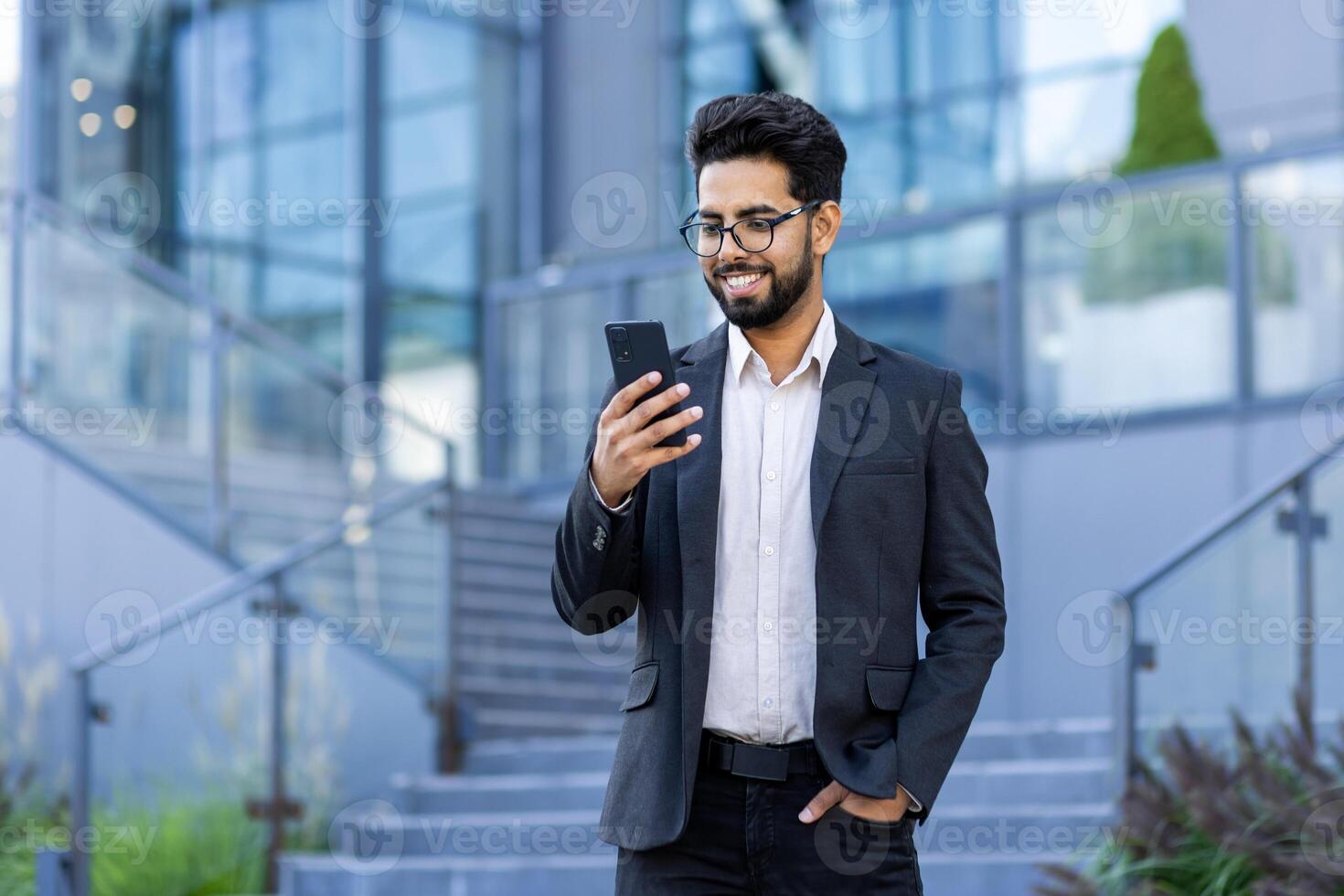 A young Indian businessman in smart casual attire is using his smartphone. Captured outside a contemporary office building, he exudes professionalism and confidence with a pleasant smile. photo