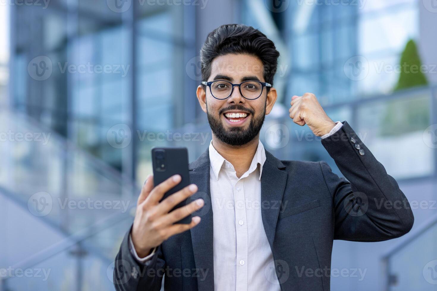 Close-up portrait of a young Muslim man in a suit and glasses standing on the street, holding a phone and showing a victory gesture with his hand. He looks at the camera with a smile. photo