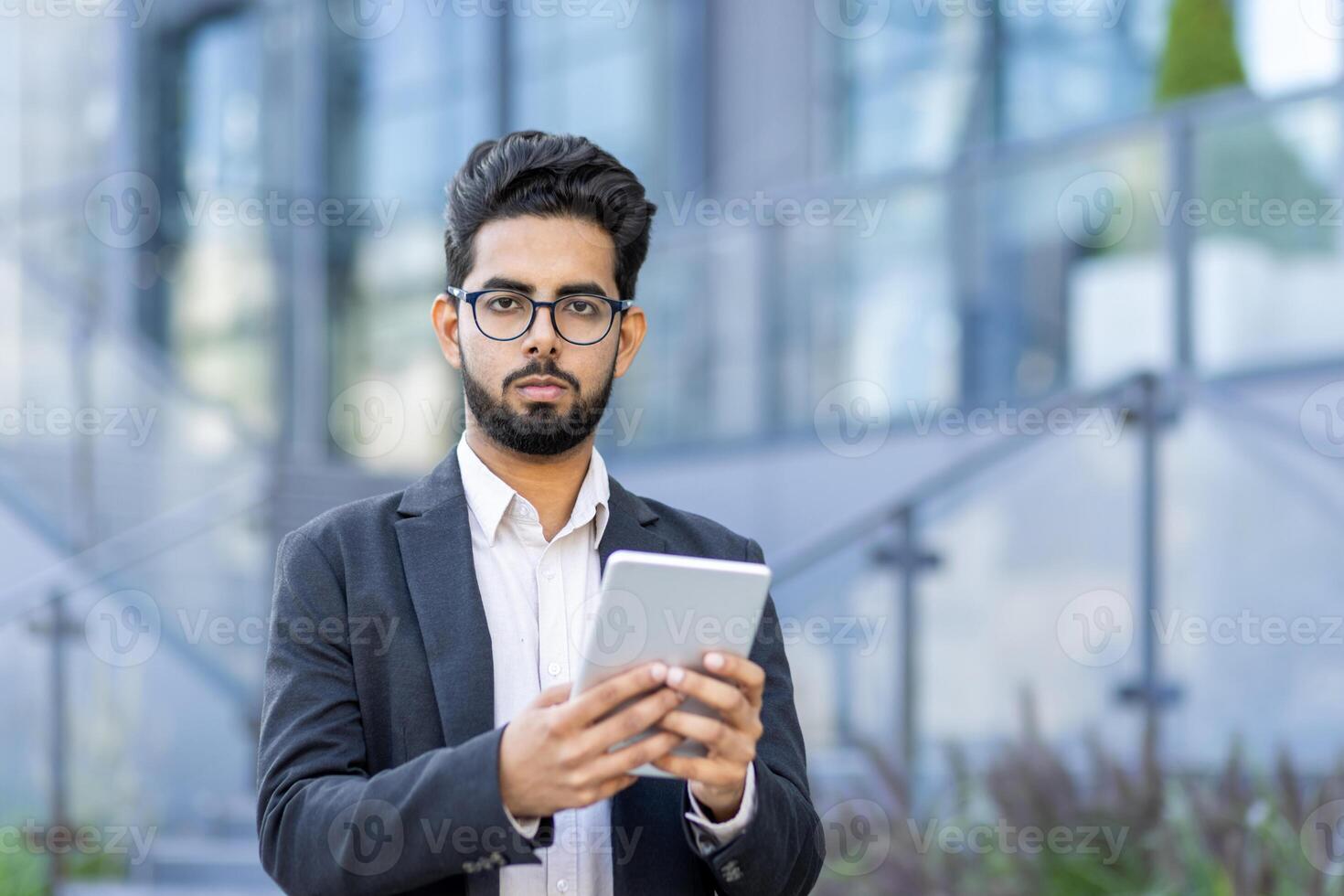 Close-up portrait of a serious young Indian man in a suit and glasses standing outside an office center, holding a tablet, looking seriously and confidently into the camera. photo