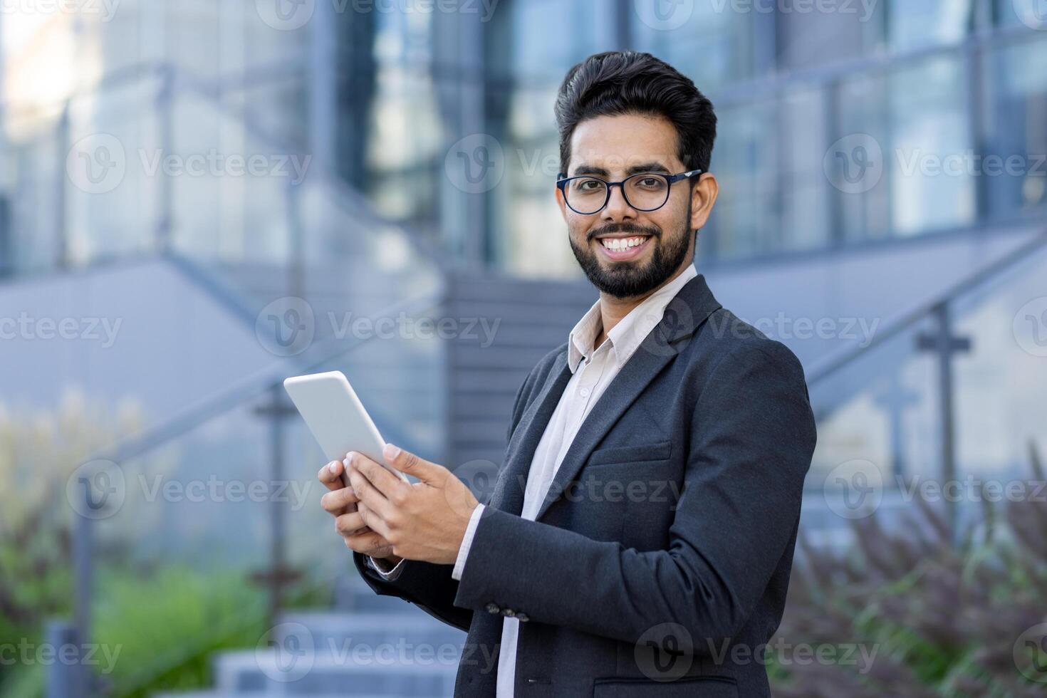 A cheerful Indian businessman in formal attire stands outdoors, using a digital tablet with a confident smile. Modern business setting with a multicultural subject. photo
