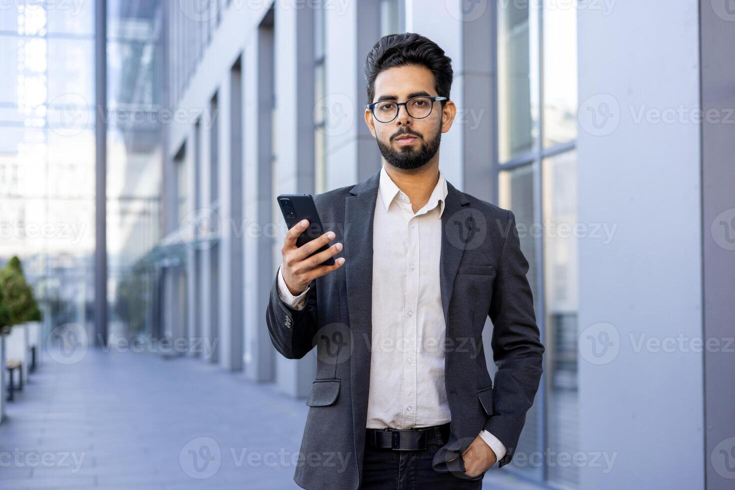 A professional Indian businessman in a smart suit stands outside modern office buildings, intently using his smartphone. This image conveys a sense of connectivity, confidence, and the fast pace of corporate life. photo
