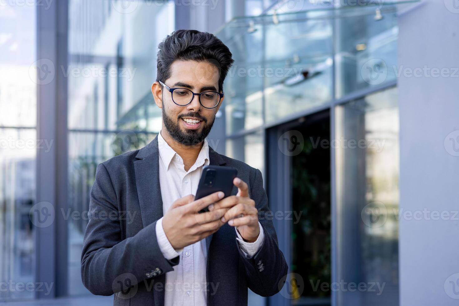 A young Indian businessman in a suit is engaged with his smartphone, smiling as he texts or reads a message. This image captures a moment of modern professional life, possibly during a break or while commuting. photo