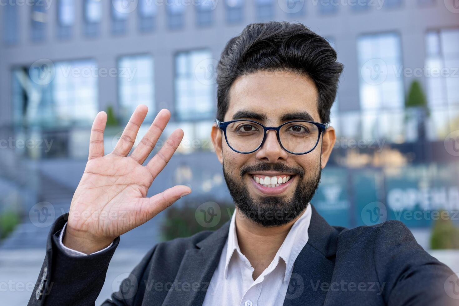 Portrait of a cheerful Indian businessman waving his hand in greeting. Standing outdoors with a bright smile, he portrays a casual yet professional demeanor. Represents multicultural business environments. photo