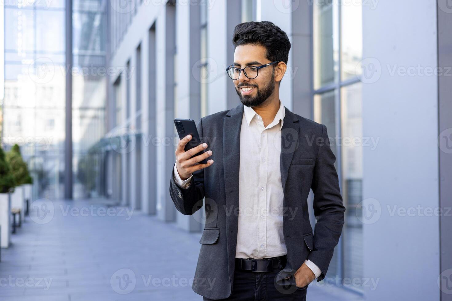 A happy and successful young Indian man in formal wear is engaged with his smartphone, smiling while standing in a modern city environment. This image captures the essence of connectivity, urban life, and professional success. photo