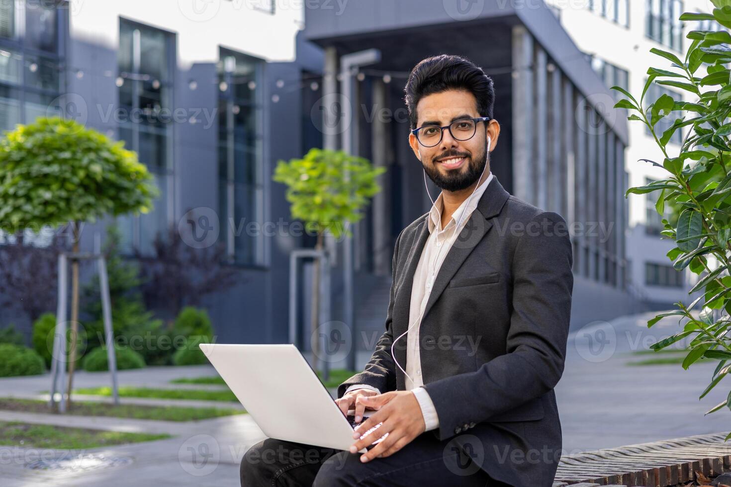 Portrait of a smiling young Indian man sitting in a business suit, wearing headphones, holding a laptop on his lap, looking confidently into the camera. photo