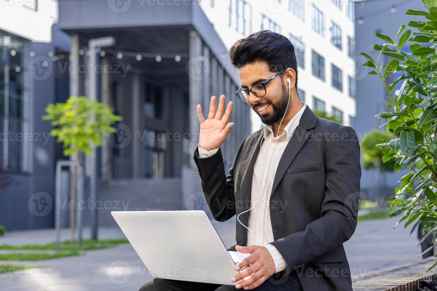 A cheerful Indian man in formal business attire is engaged in a call using his laptop outdoors, conveying a sense of connectivity, professionalism, and modern business practices in a casual outdoor environment. photo