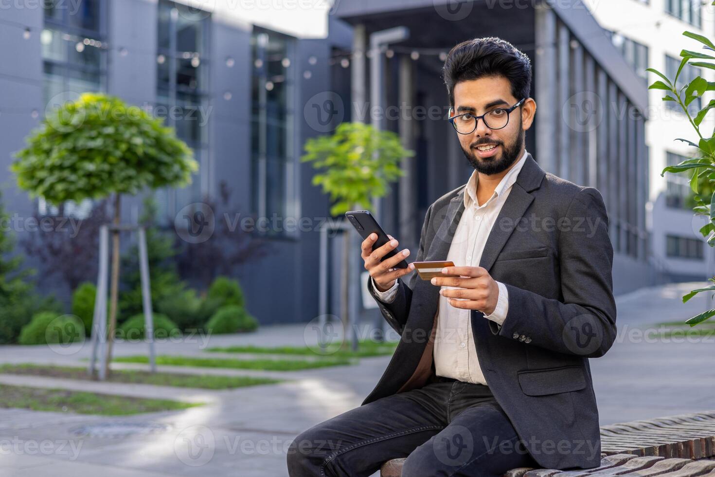 Portrait of a young Indian man in a business suit sitting on a bench near an office center, holding a credit card and a phone, looking at the camera with a smile. photo