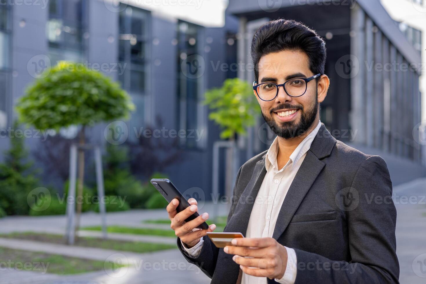 Close-up portrait of a young Indian businessman man sitting in a suit outside on a bench, holding a credit card and a mobile phone, smiling and looking at the camera. photo