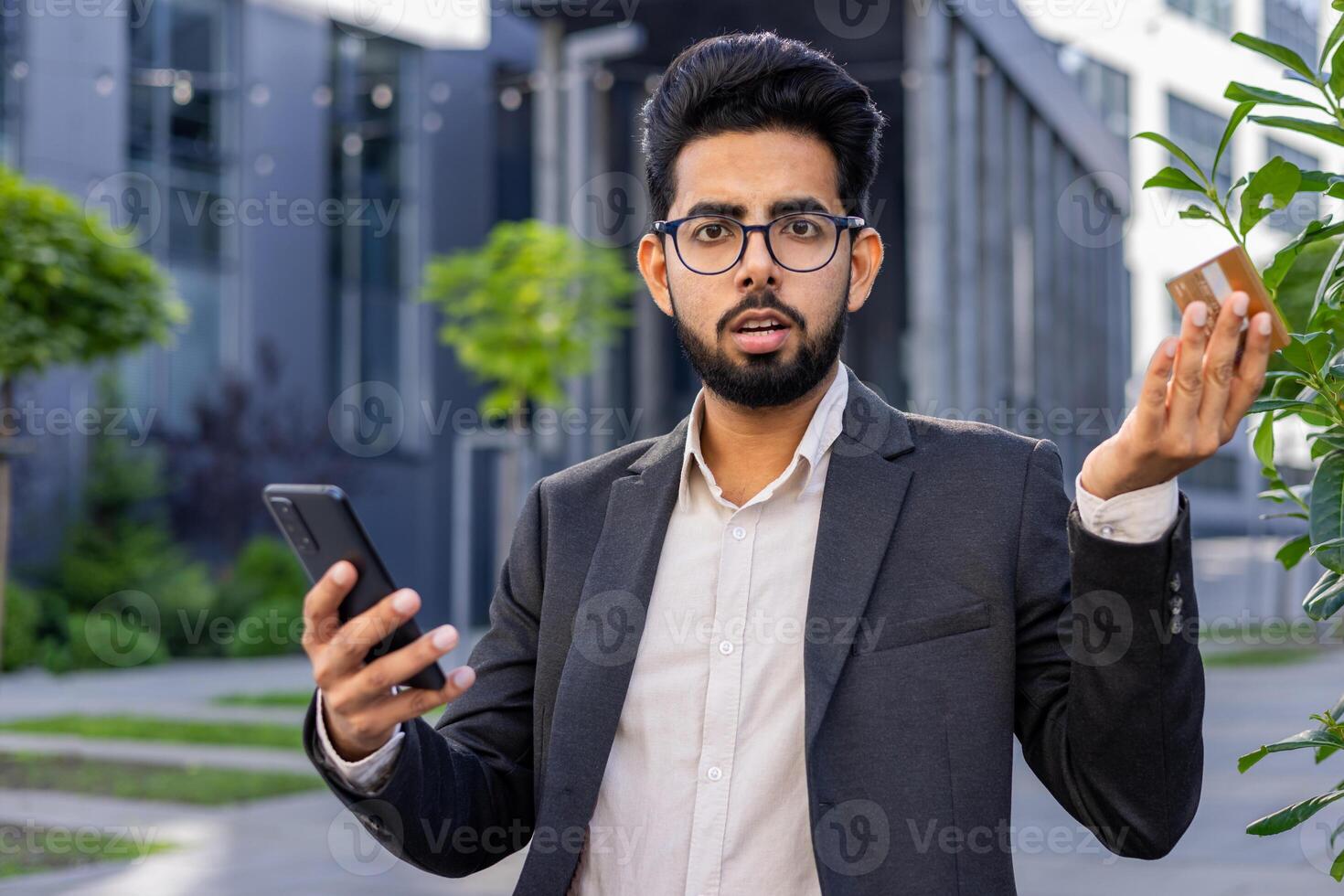 Portrait of a worried young Indian male businessman in a business suit sitting on a bench near the office, holding a credit card and a phone, looking disappointedly at the camera and spreading hands. photo
