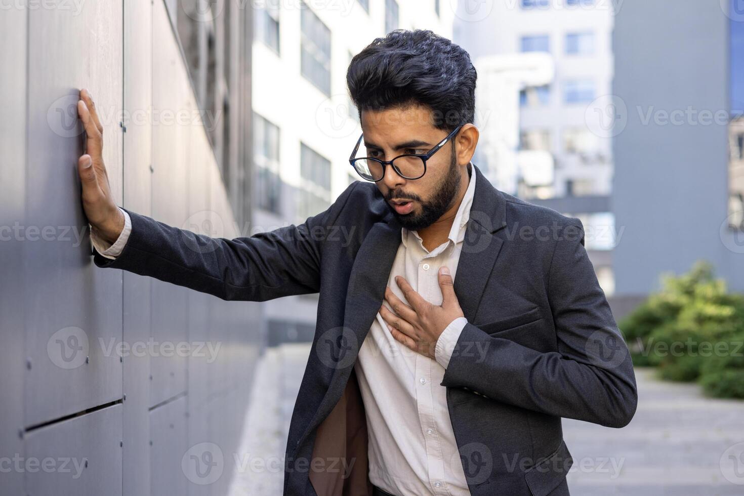 A professional man in a business suit pauses for a moment outside, resting against a wall with a hand over his heart, suggesting contemplation, sincerity, or a health concept. photo