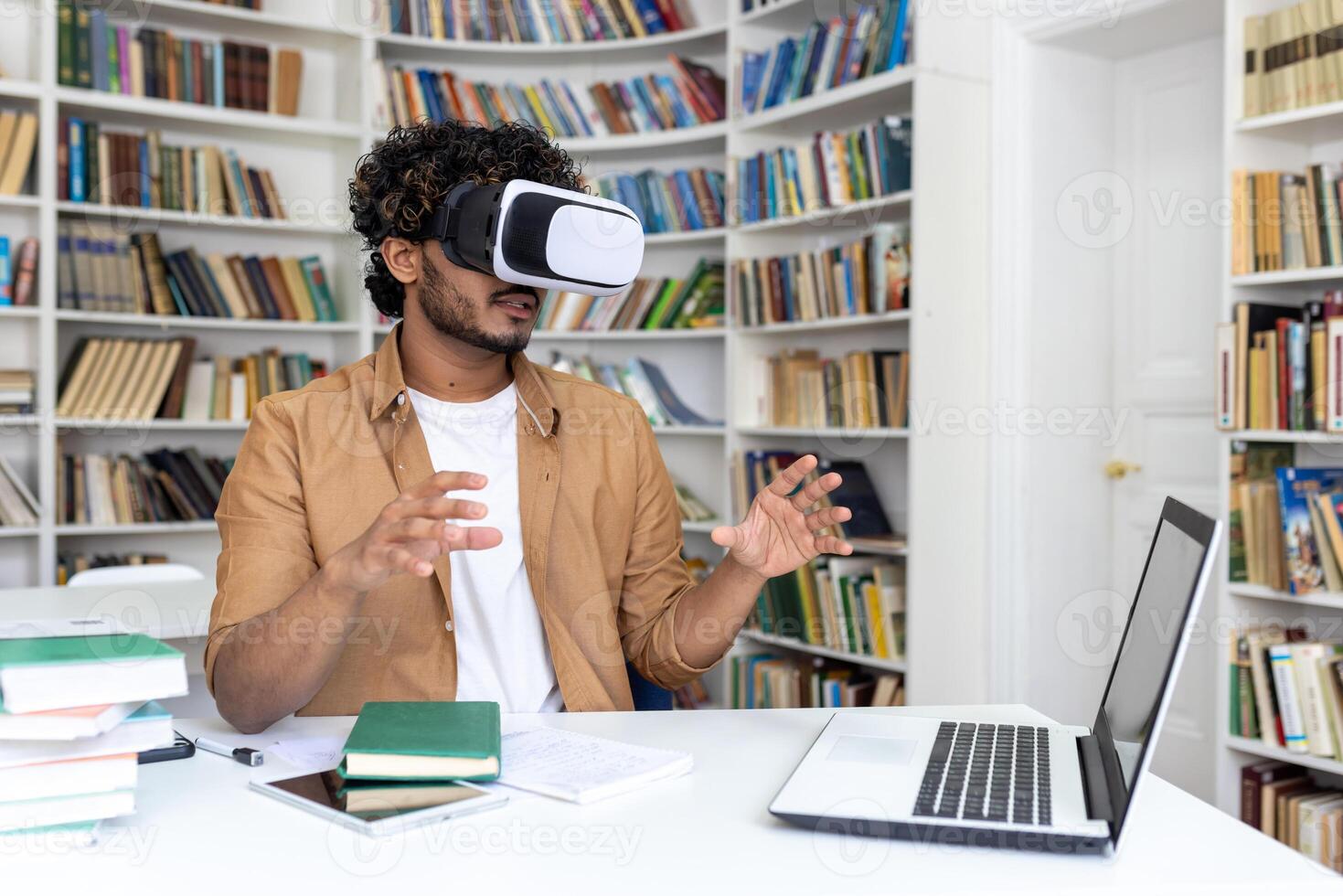 Young hispanic male student sitting at a desk in a university library, studying online on a laptop wearing a virtual mask. Gesturing and waving his hands. photo