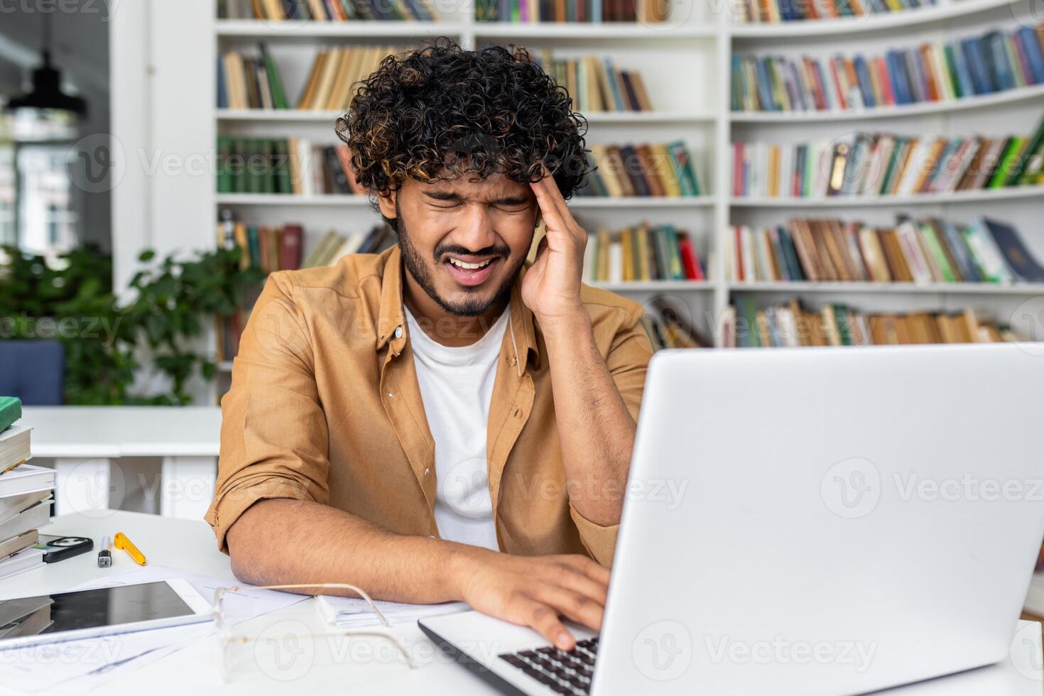 A young man with curly hair is visibly stressed and experiencing a headache as he works on his laptop, possibly dealing with work pressure or studying challenges in a home environment photo