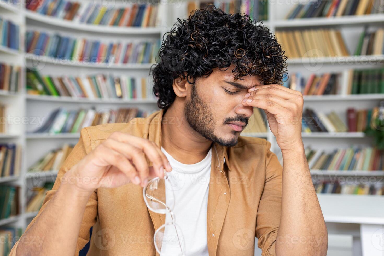 A young man with curly hair appears stressed and holds his head in pain, showing signs of a headache. He has taken off his eyeglasses, perhaps after reading or studying for long hours. photo
