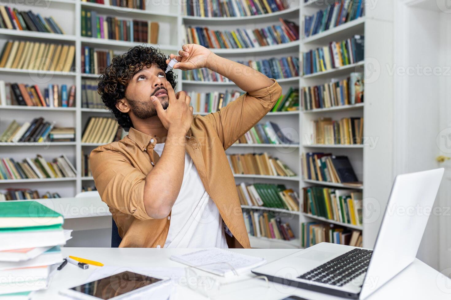 Concentrated islamic man with curly hair throwing head back while using eye drops and sitting in cabinet. Responsible male using prevention medicine for avoiding vision problems after work with pc. photo