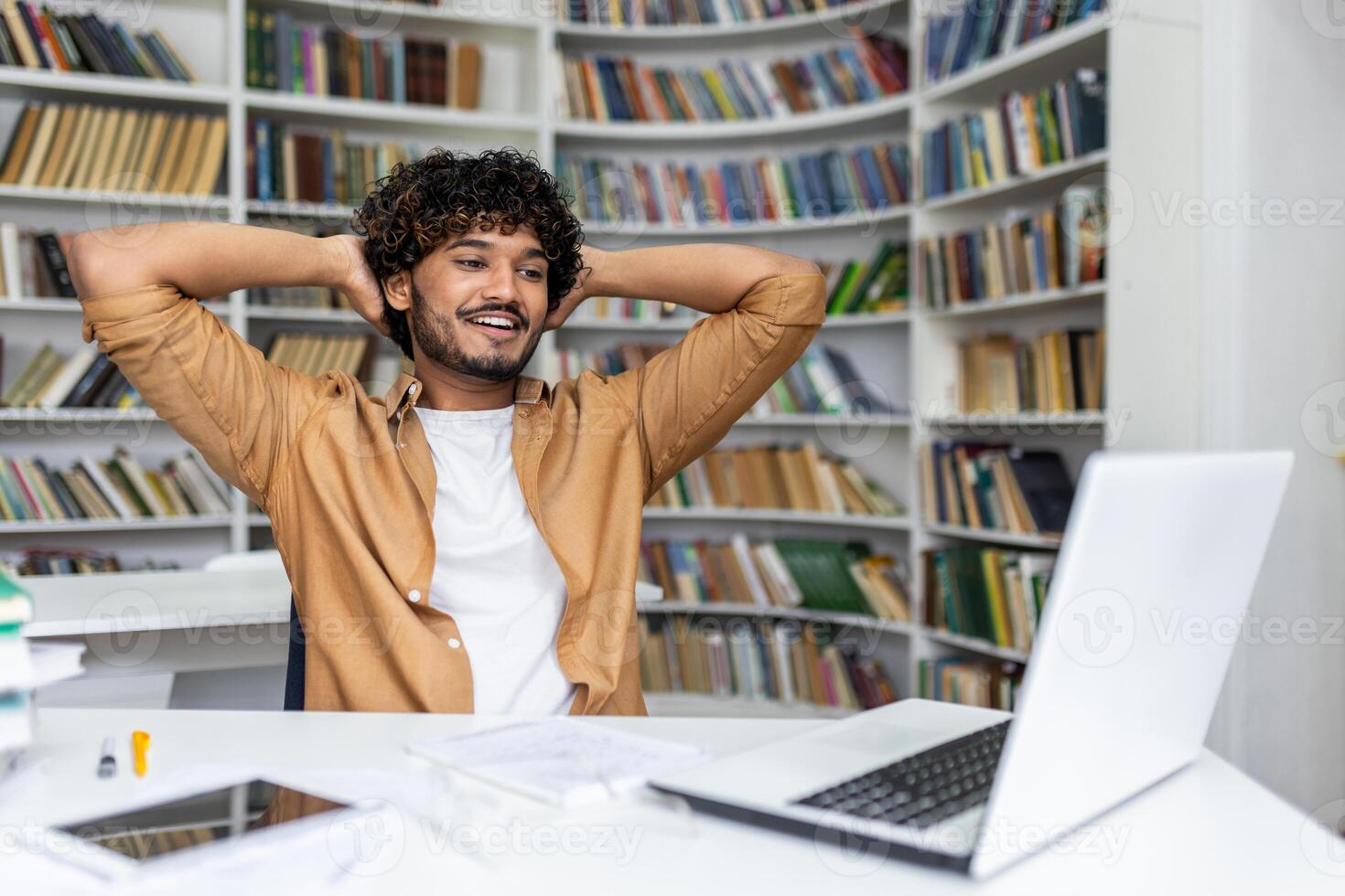 Positive hispanic guy leaning on back of chair while holding hands behind head and looking at laptop screen. Smiling student feeling satisfied after passing exam remotely and getting high grade. photo
