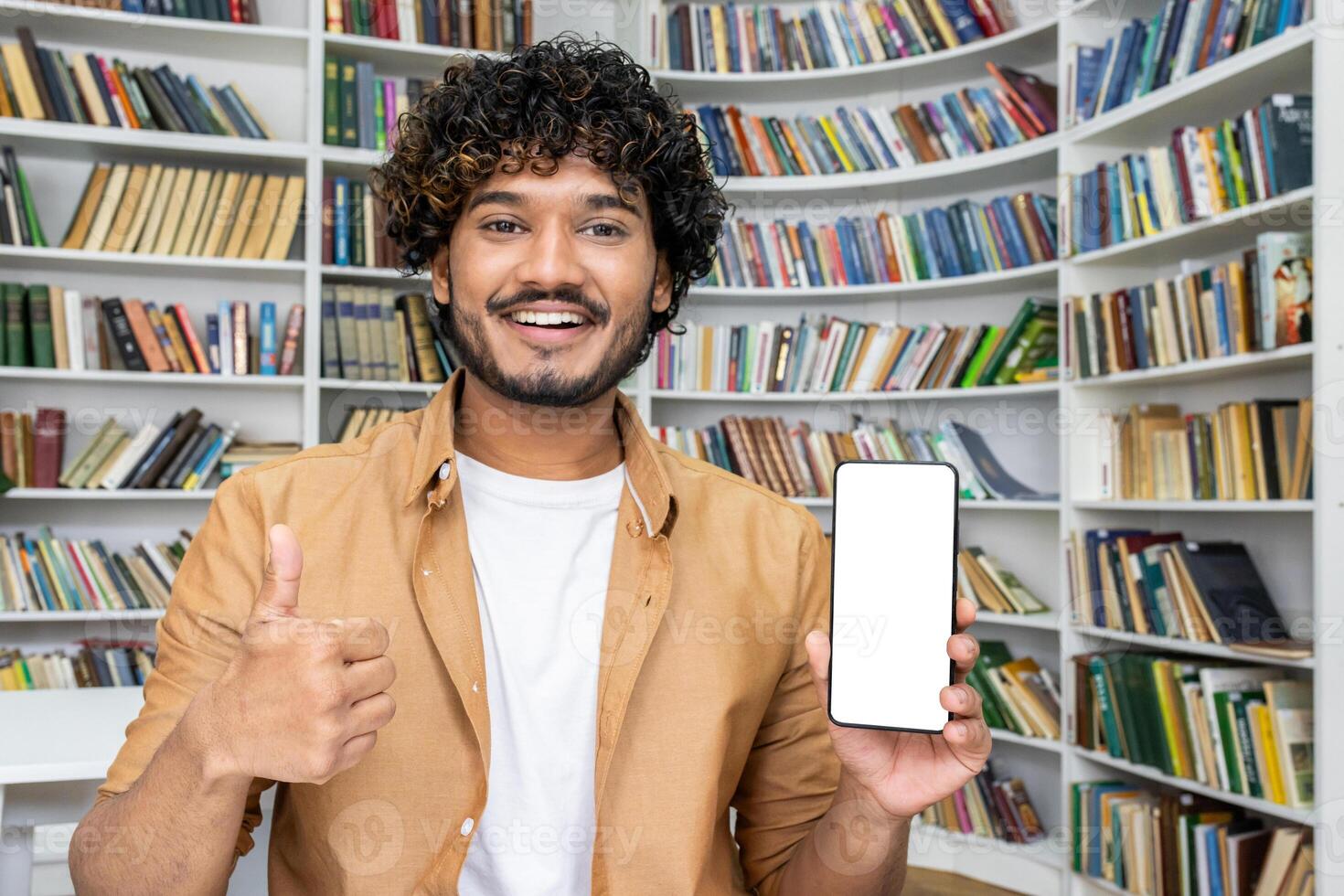 A young man stands in front of a bookshelf in a library, presenting a smartphone with a blank screen, giving a thumbs-up sign, suggesting approval or a positive review of an app. photo