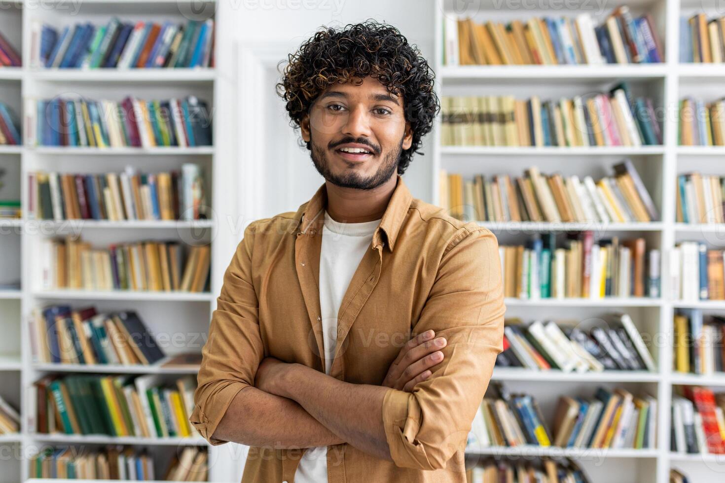 Confident diverse man in casual outfit looking at camera with arms folded on chest on background of closet with books. Intelligent male posing in library of university campus with happy expression. photo
