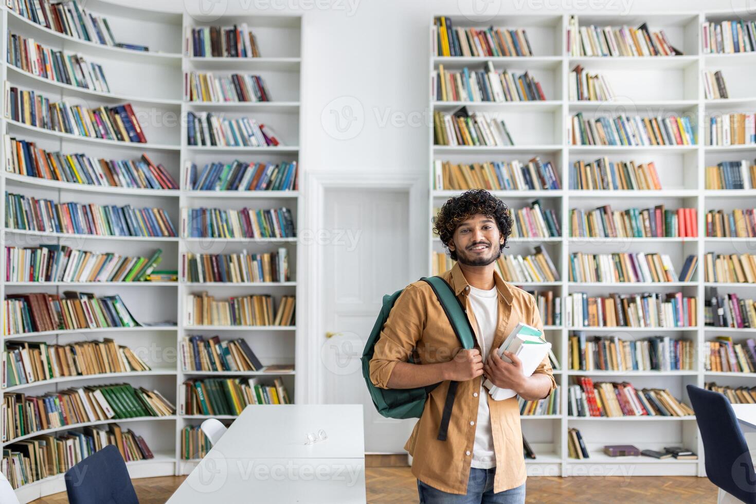 A cheerful young adult with curly hair and a backpack standing in a bright library, holding several books, exuding a vibe of learning and positivity amid shelves full of books. photo