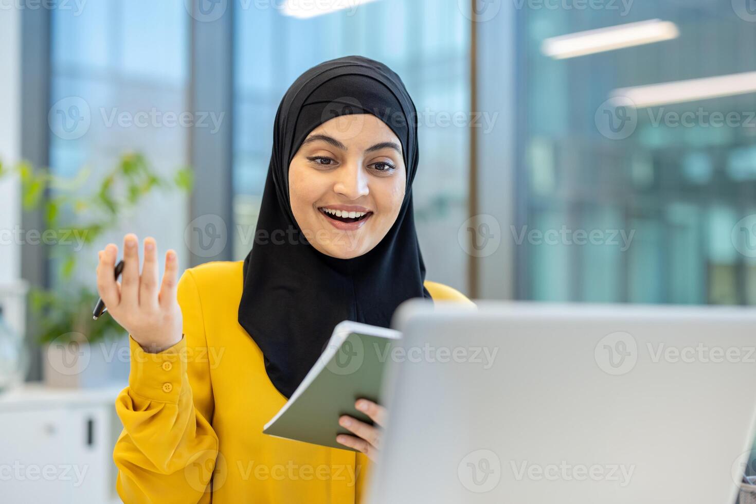 A young woman in a hijab smiles broadly while looking at a laptop, holding a notebook and pen in an office setting. photo