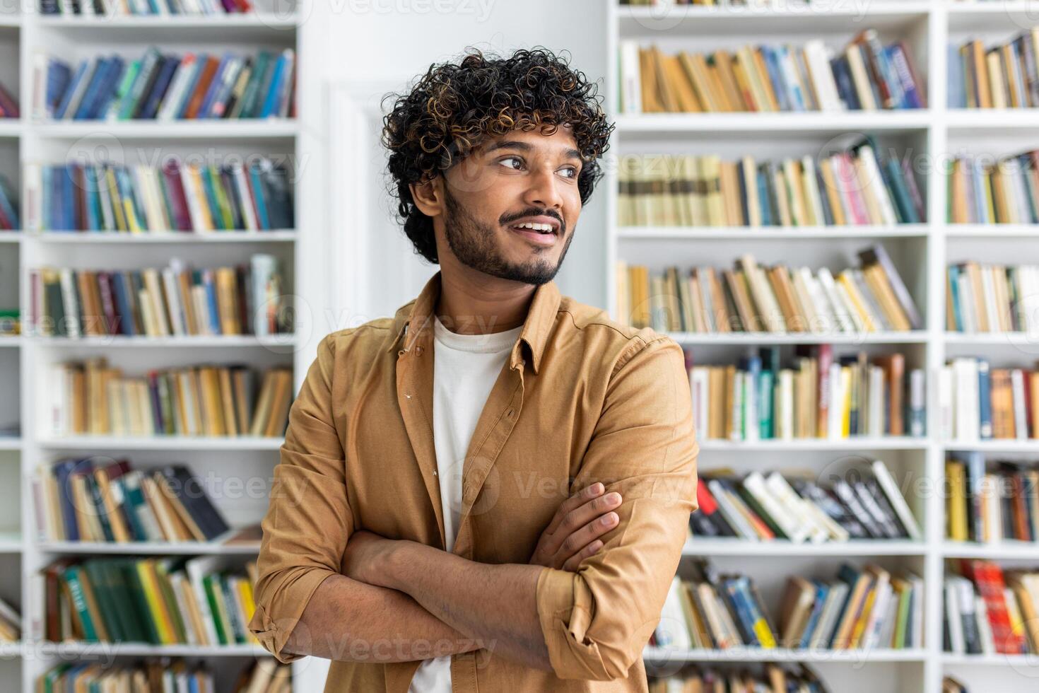 A casually dressed professional stands assuredly with arms crossed in front of a bookshelf. The focus is on confidence and casual professionalism within an educational or literary setting. photo