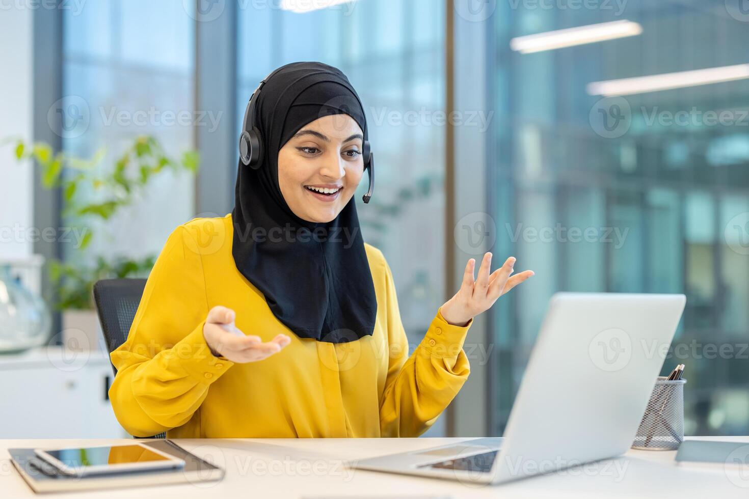 A smiling woman in a yellow shirt and hijab speaks into a headset while looking at her laptop in an office setting. photo