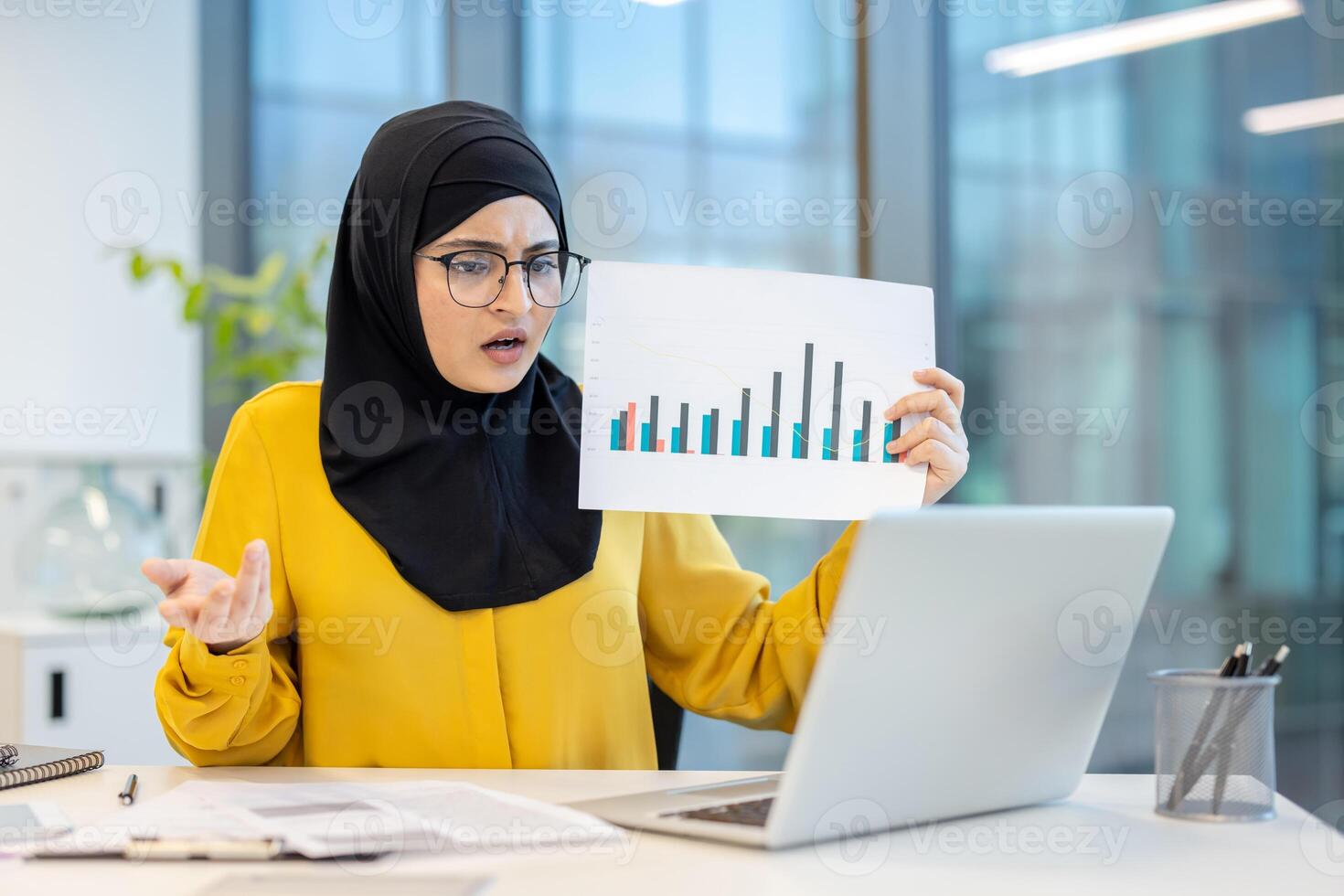 A woman wearing a hijab is looking at a financial chart on a laptop, appearing concerned about the data. photo