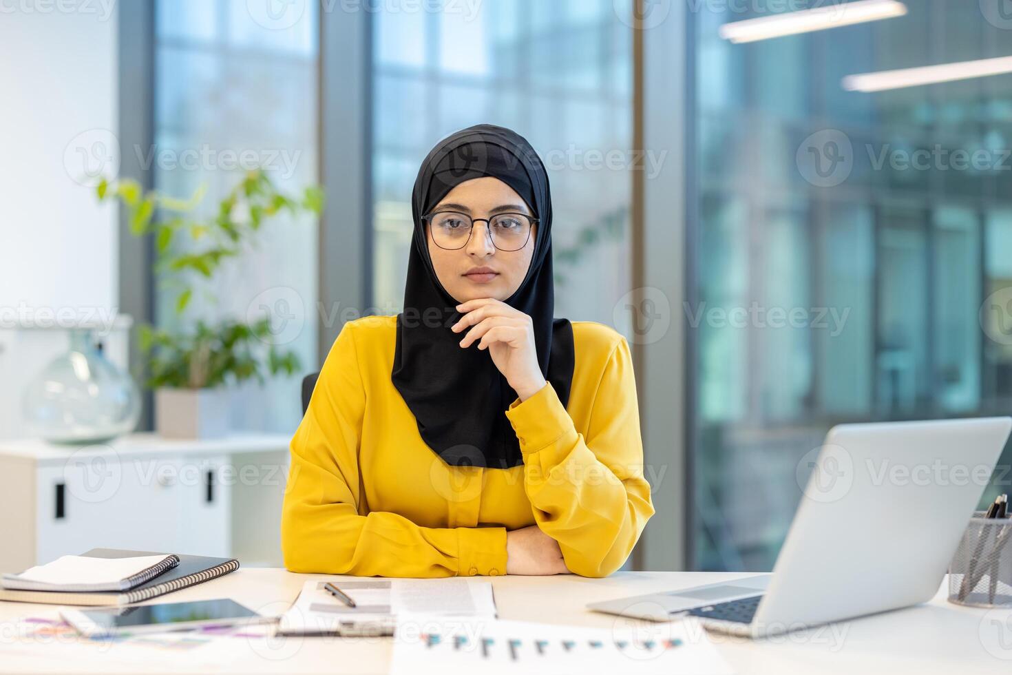 A Muslim businesswoman wearing a hijab sits at her desk in an office, looking at the camera with a serious expression. photo