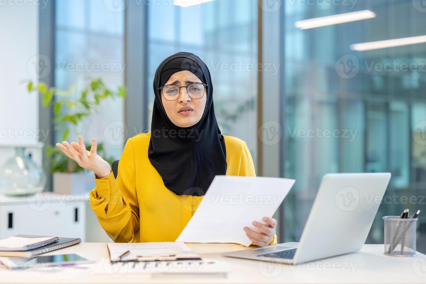A surprised Muslim woman in glasses looks at documents in an office, surrounded by work tools. photo