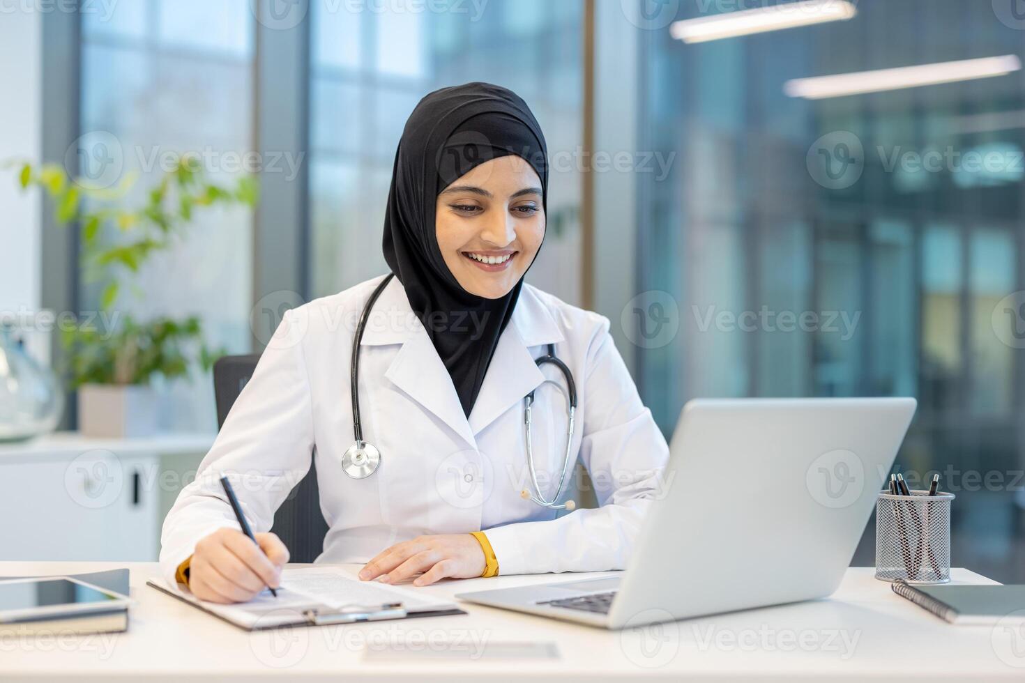 A smiling female doctor, wearing a hijab, works at a desk with a laptop, taking notes, in a modern office setting. photo
